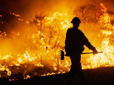 A firefighter holding a rake walks toward the flames surrounding him