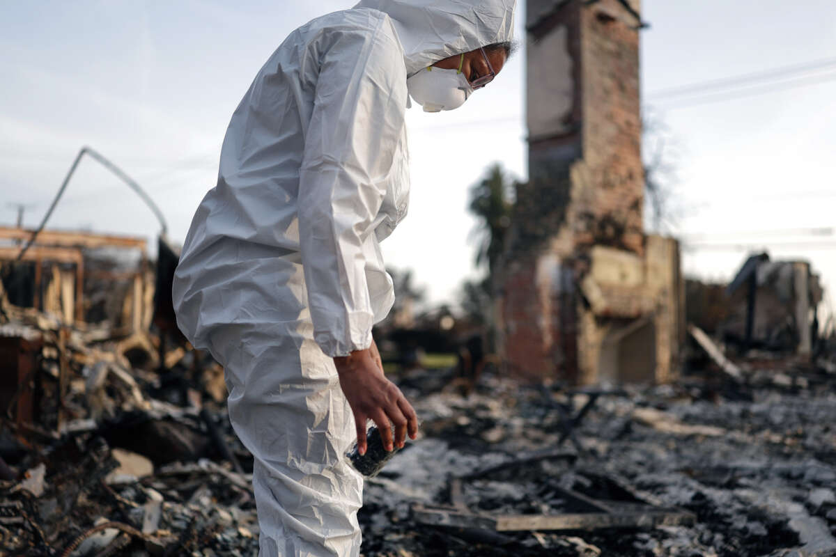 A person in a hazmat suit collects ashes from a destroyed building into a jar