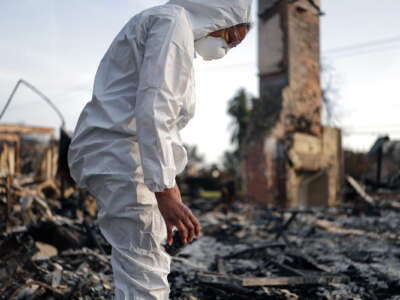 A person in a hazmat suit collects ashes from a destroyed building into a jar