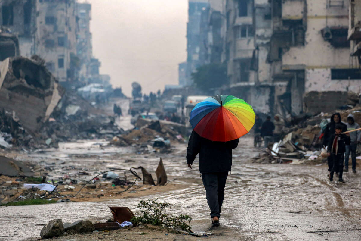 A man walks with a rainbow-colored umbrella along a muddied street past the rubble of destroyed and heavily damaged buildings in the north of Gaza City on February 10, 2025.