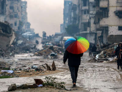 A man walks with a rainbow-colored umbrella along a muddied street past the rubble of destroyed and heavily damaged buildings in the north of Gaza City on February 10, 2025.