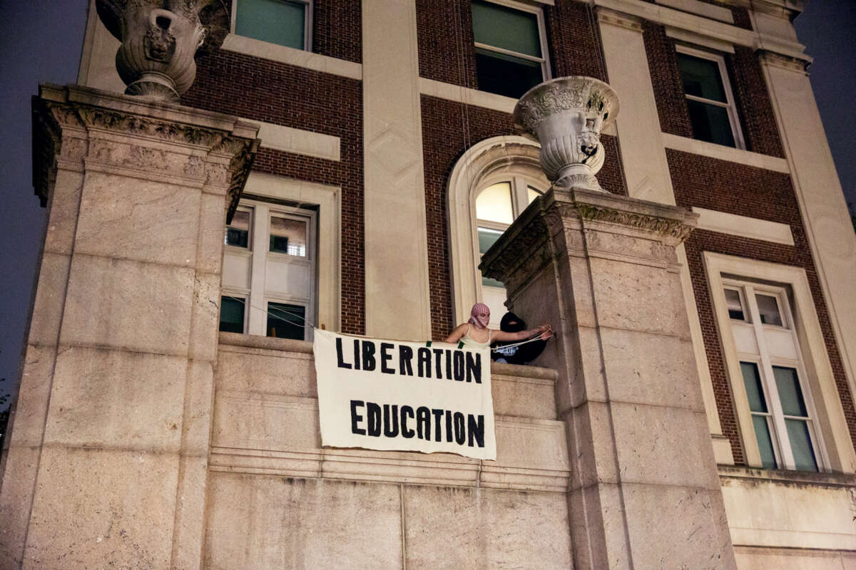 Two students unfurl a banner reading "LIBERATION; EDUCATION" at a university building