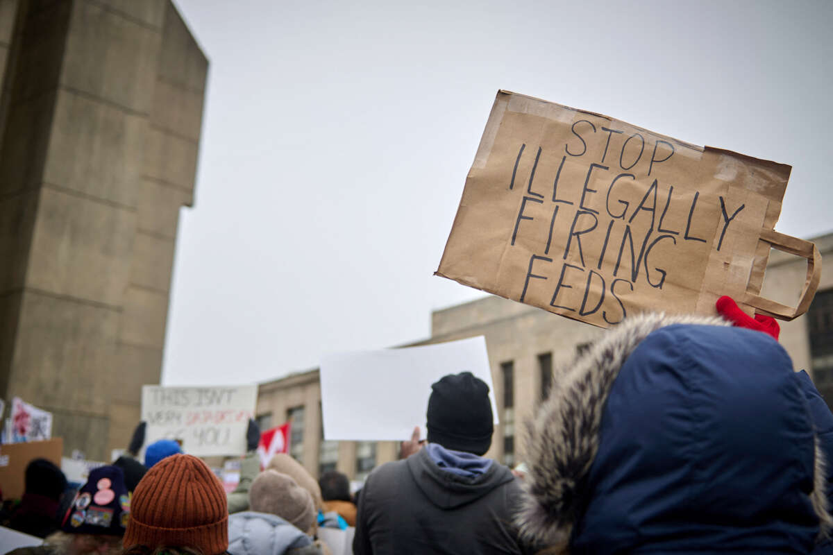 A protester holds a sign reading "STOP ILLEGALLY FIRING FEDS" during an outdoor demonstration