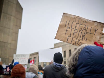 A protester holds a sign reading "STOP ILLEGALLY FIRING FEDS" during an outdoor demonstration