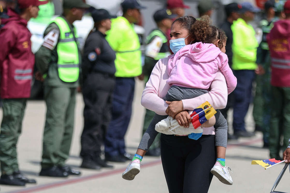 A woman wearing a medical face mask carries her daughter in one hand and a tiny Venezuelan flag in the other