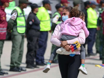 A woman wearing a medical face mask carries her daughter in one hand and a tiny Venezuelan flag in the other