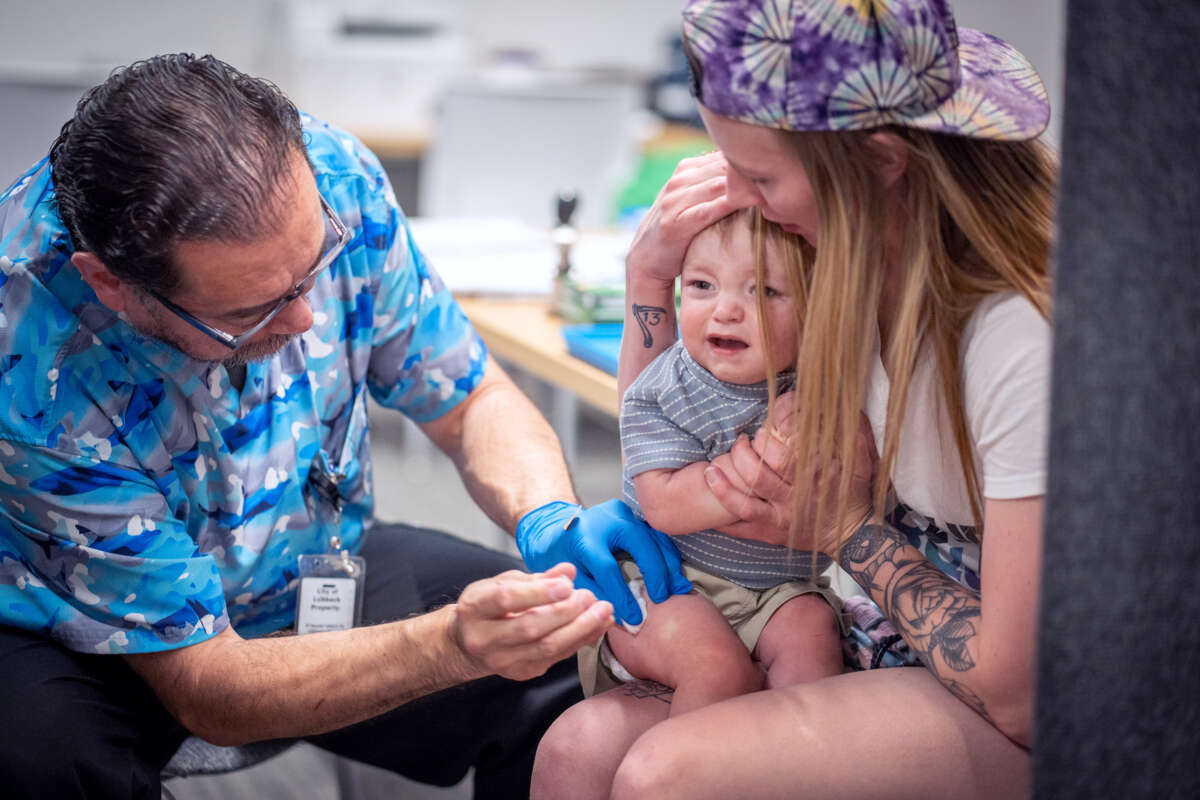 A medical worker in a brightly-patterned blue shirt administers the mmr vaccine to a crying child being held by his mother