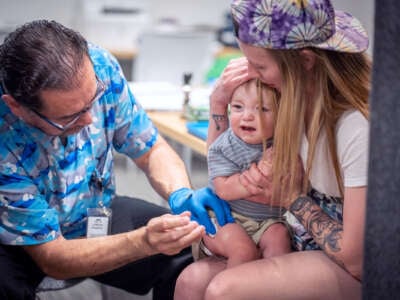 A medical worker in a brightly-patterned blue shirt administers the mmr vaccine to a crying child being held by his mother