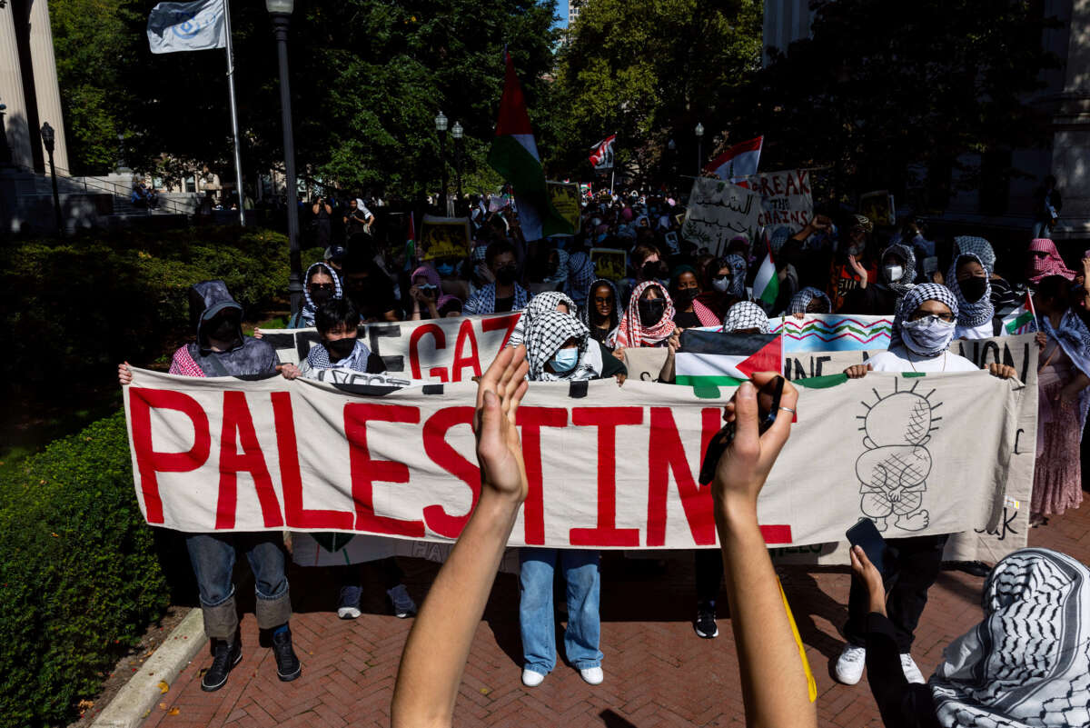 Students with covered faces march behind a banner that reads "PALESTINE"