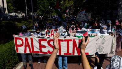 Students with covered faces march behind a banner that reads "PALESTINE"