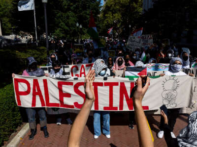 Students with covered faces march behind a banner that reads "PALESTINE"