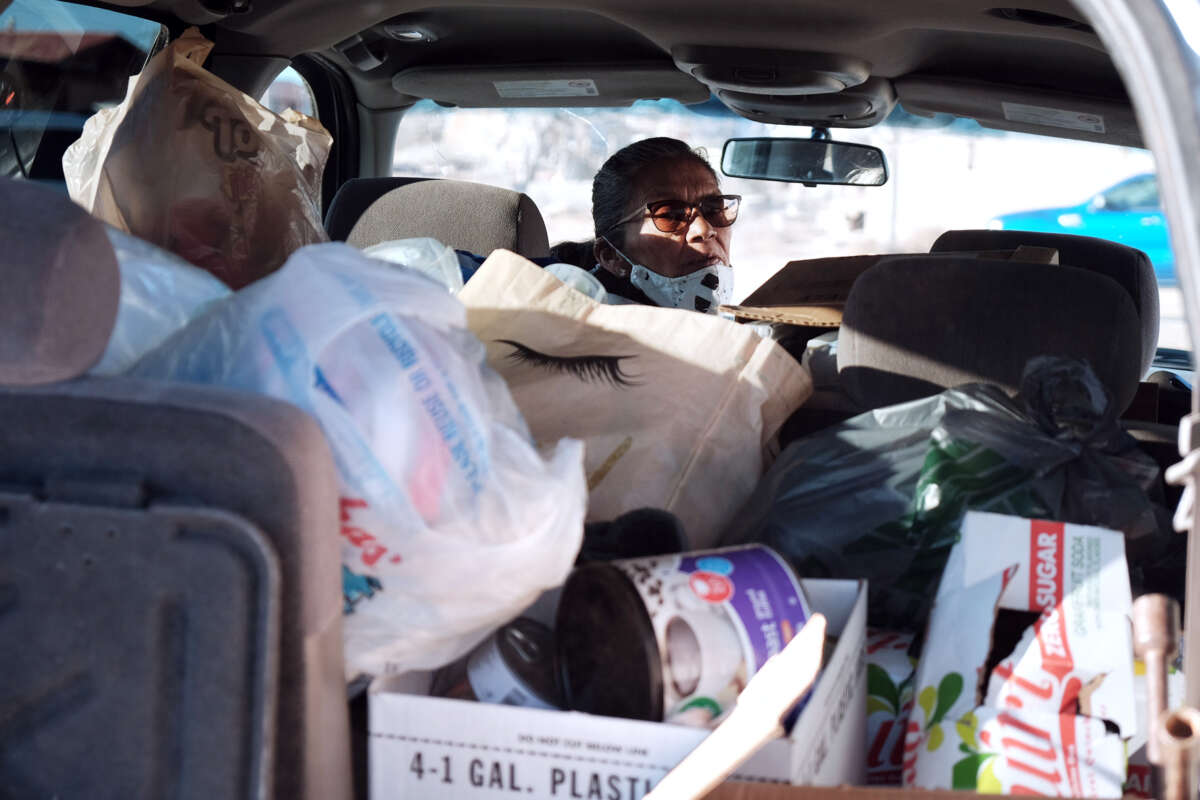 A woman wearing a mask looks behind herself from the drivers seat of the van she's currently in, which is filled with goods