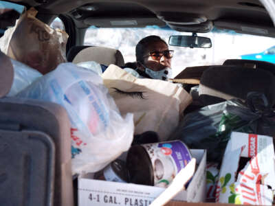 A woman wearing a mask looks behind herself from the drivers seat of the van she's currently in, which is filled with goods