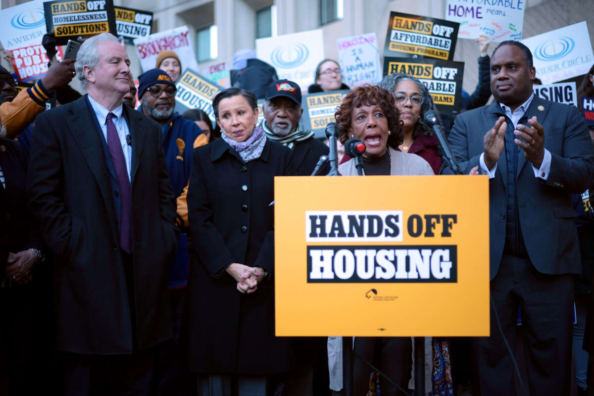 Rep Barbara Lee, surrounded by others, speaks at a podium with an affixed sign reading "HANDS OFF HOUSING"