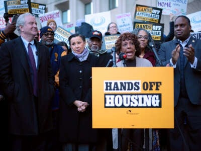 Rep Barbara Lee, surrounded by others, speaks at a podium with an affixed sign reading "HANDS OFF HOUSING"