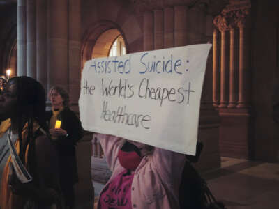 A person holds a sign that reads, ‘ASSISTED SUICIDE: The World’s Cheapest Healthcare.’ They are a wheelchair user with two face masks on, wearing a pink shirt that says ‘NOT DEAD YET,’ in the middle of a busy room at a protest at the New York State Assembly.