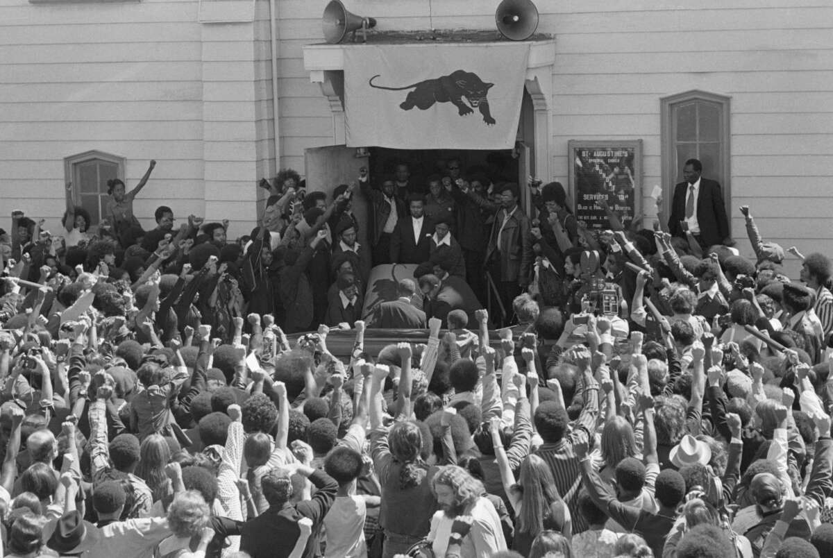 A black and white image of mourners giving the Black Panther salute as the casket of George Jackson is carried from St. Augustine's Church in Oakland, California.