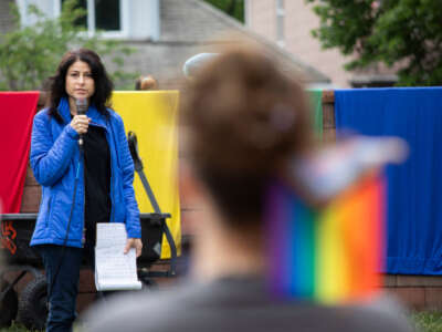 AG Dana Nessel is on a platform holding a mic and addressing a crowd of protesters. Around her are flags including the LGBT Rights flag.
