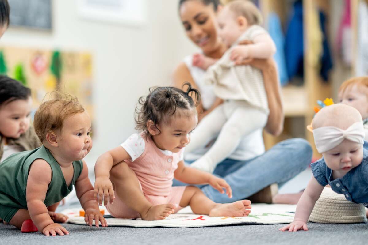 A group of babies is seen playing on the floor of their daycare classroom