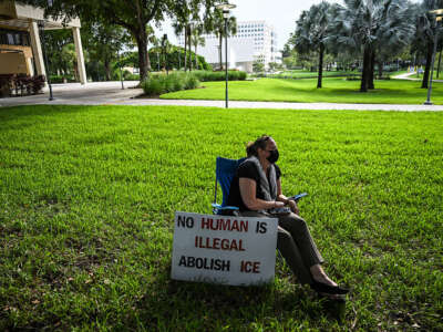 Woman sits on a lawn chair with a sign next to her. She is also wearing a face mask.