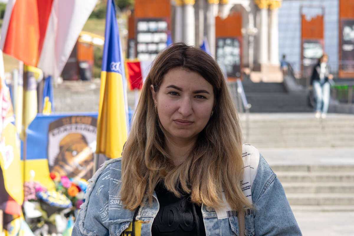 A woman with blonde hair stands in front of a Ukrainian flag.