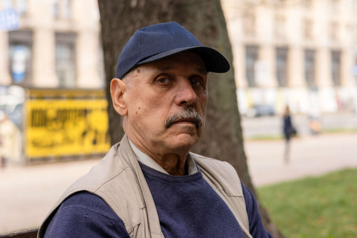 A man with a blue hat and mustache stands in front of a tree.