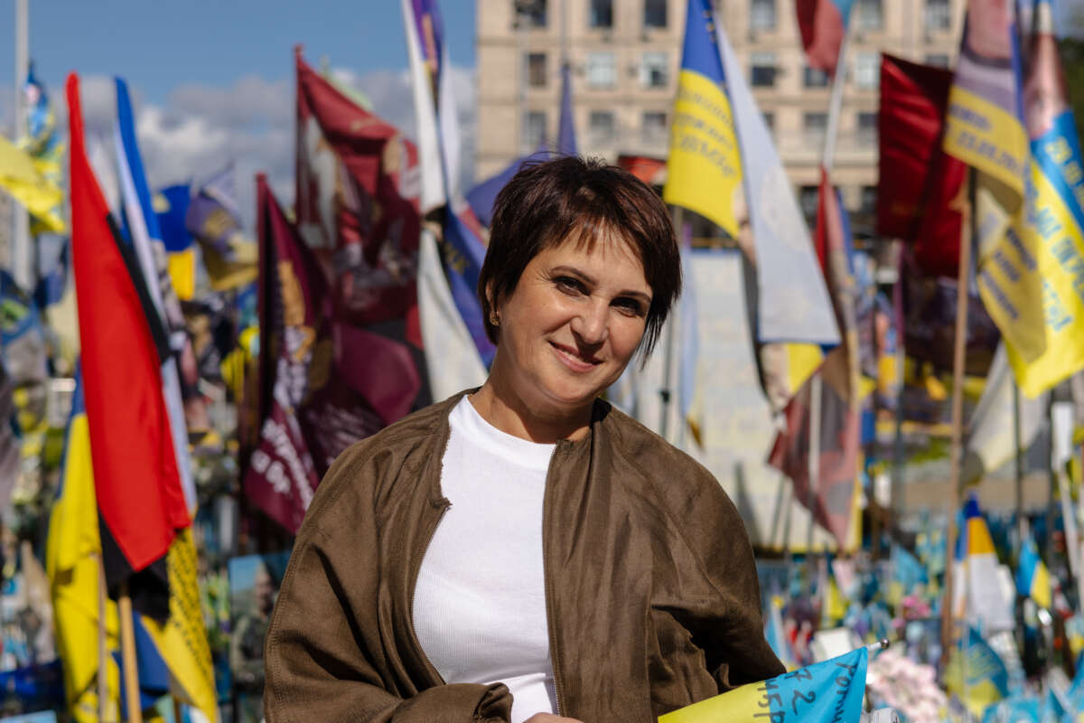 A woman with short hair and a brown jacket stands in front of a variety of flags.