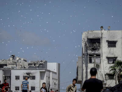 People are seen at the bottom of the picture picking up flyers that are coming down. The sky is filled with many of them still coming down.