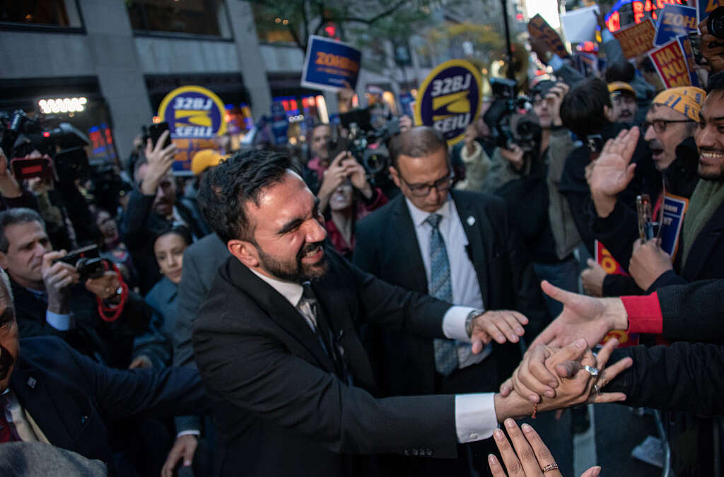 Zohran Mamdani is seen shaking hands with people as other people rally behind him with signs.