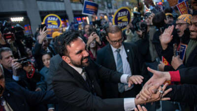 Zohran Mamdani is seen shaking hands with people as other people rally behind him with signs.
