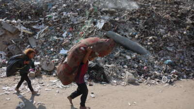 Palestinian children carry their belongings past an unexploded missile in a rubbish dump at the Firas market area of Gaza City, on November 12, 2025.
