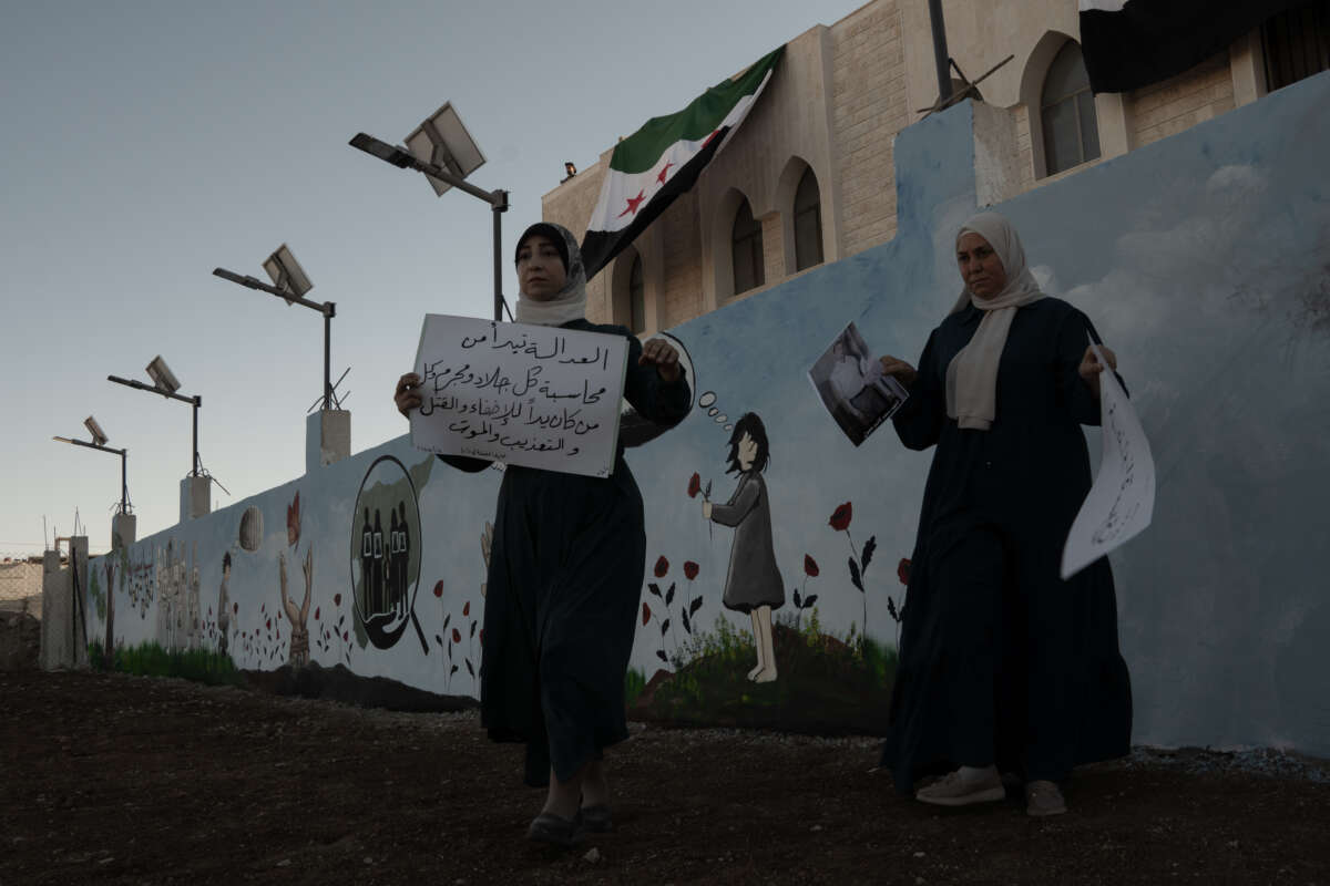 Two women hold signs while standing in front of a wall painted with a child in a field of wildflowers.