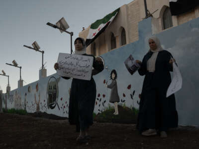 Two women hold signs while standing in front of a wall painted with a child in a field of wildflowers.