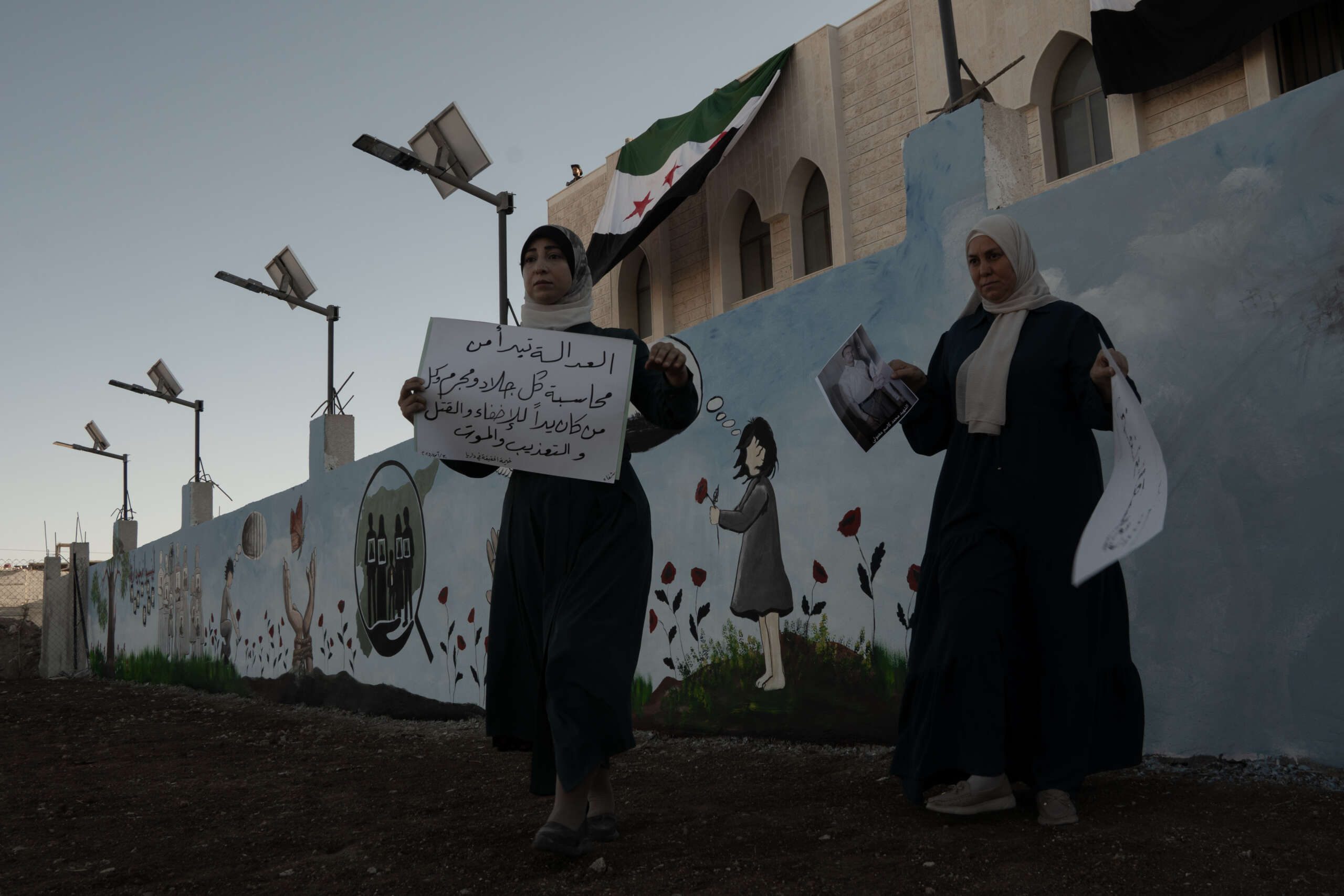 Two women hold signs while standing in front of a wall painted with a child in a field of wildflowers.