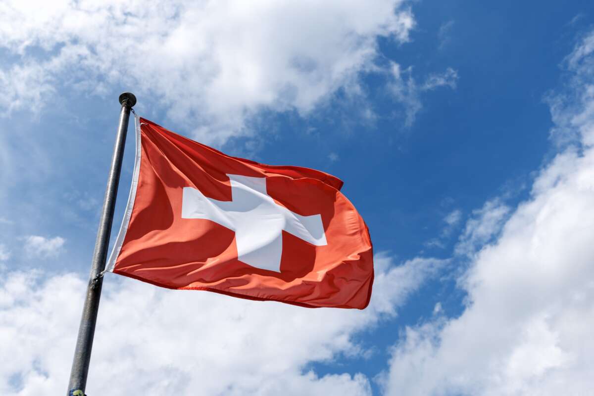A rectangular flag of Switzerland flies in the wind on a flagpole against the blue sky.