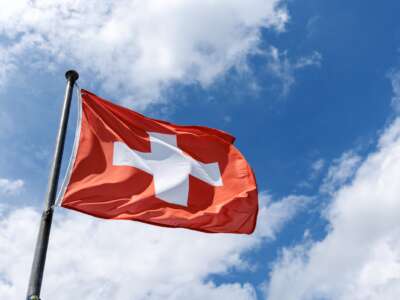A rectangular flag of Switzerland flies in the wind on a flagpole against the blue sky.