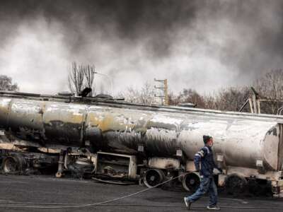 An Iranian civil defence member walks with a hose next to a destroyed fuel tanker vehicle near an ongoing fire following an overnight airstrike on the Shahran oil refinery in northwestern Tehran on March 8, 2026.