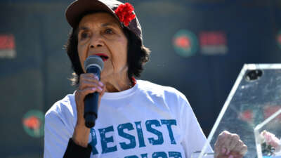 Dolores Huerta holding a microphone and wearing a shirt that reads "resist"