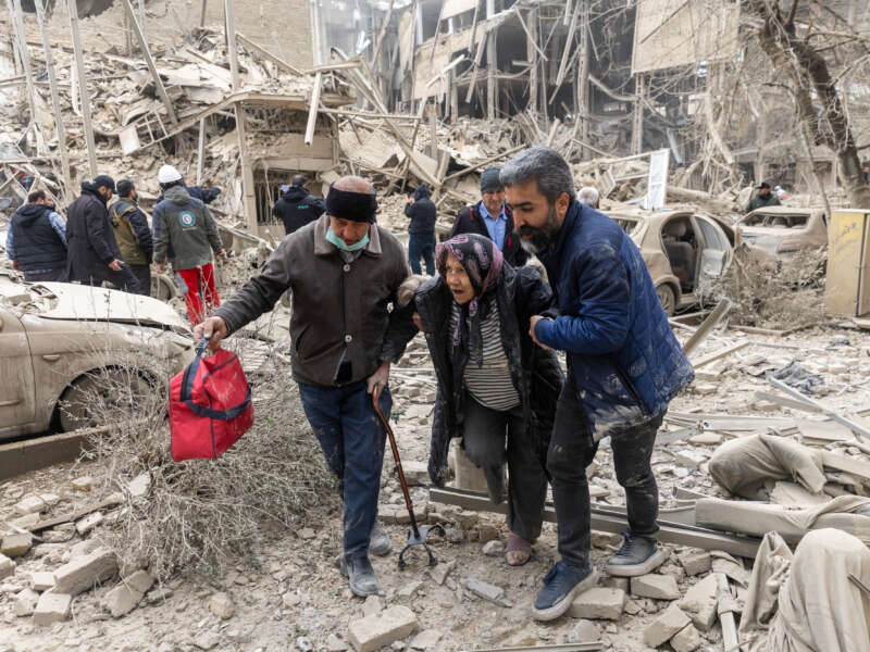 An elderly woman is helped from the scene of a strike on a residential building on March 16, 2026, in Tehran, Iran.