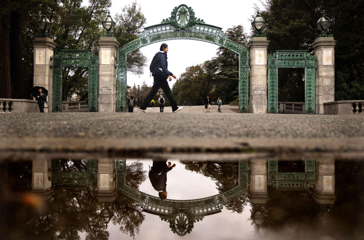 A student is reflected in a rain puddle while passing by Sather Gate at the University of California, Berkeley, on February 19, 2026.