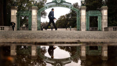 A student is reflected in a rain puddle while passing by Sather Gate at the University of California, Berkeley, on February 19, 2026.