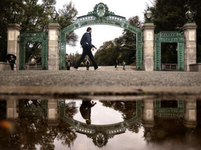 A student is reflected in a rain puddle while passing by Sather Gate at the University of California, Berkeley, on February 19, 2026.