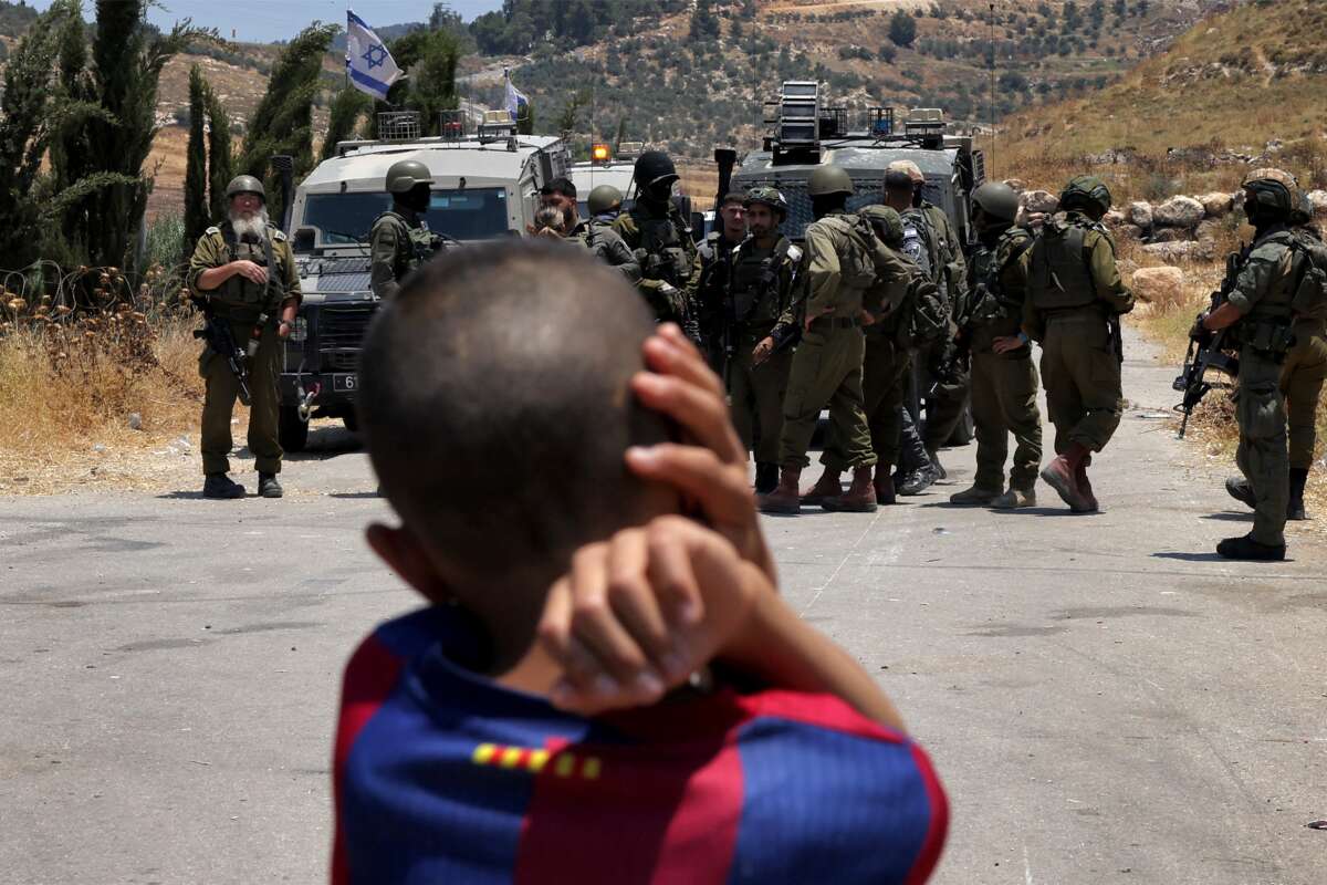 A Palestinian boy looks on as Israeli forces stand guard before Palestinians gathering to protest threats of confiscation of their land in the Palestinian village of Idna, west of Hebron in the occupied West Bank, on June 12, 2026.