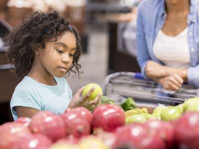 A serious elementary age little girl stands in the produce section of a grocery store and looks down at a granny smith apple in concentration as she inspects it. Her unrecognizable mother waits patiently by the cart.