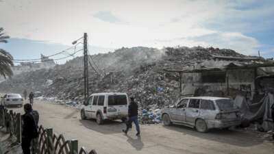 People walk and drive along a road beside large piles of debris at the destroyed Firas Market in Gaza City, on December 9, 2025. The public market, one of Gaza City's oldest and busiest commercial hubs, has been left buried under rubble and waste after widespread destruction from Israeli strikes and the collapse of basic municipal services.