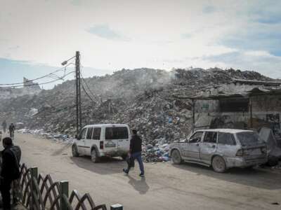 People walk and drive along a road beside large piles of debris at the destroyed Firas Market in Gaza City, on December 9, 2025. The public market, one of Gaza City's oldest and busiest commercial hubs, has been left buried under rubble and waste after widespread destruction from Israeli strikes and the collapse of basic municipal services.
