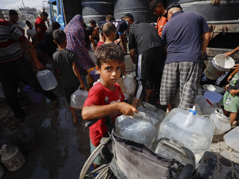 A Palestinian child stands next to a wheelchair loaded with water containers near a water distribution truck in Al-Shati refugee camp, west of Gaza City, Gaza Strip, on July 4, 2026.