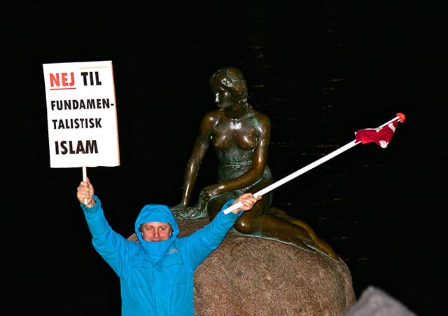 An enthusiastic PEGIDAdk supporter waves the Danish flag and a “No to fundamentalist Islam” sign in front of the Little Mermaid statue. (Photo: Linda Pershing)