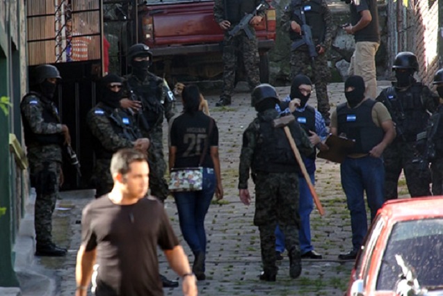 Outside Edwin Espinal’s home (Espinal is in the foreground), public prosecutor Ricardo Adolfo Núñez, wearing a blue plaid shirt, balaclava and bulletproof vest, confers with members of the military police in charge of the raid. One carries on his back a large mallet used in the operation. (Photo: Jesse Freeston)
