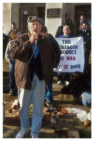 Phil Berrigan in front of a wreckage of war die-in at the River Entrance. Frida is half-visible behind to the left. Photo taken by Rick Reinhard on December 30, 1996. The banner reads, The Pentagon Product Is War: Shut It Down.
